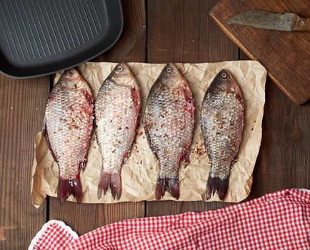 fresh crucian fish sprinkled with spices lies on brown crumpled paper, wooden table from boards, top view
