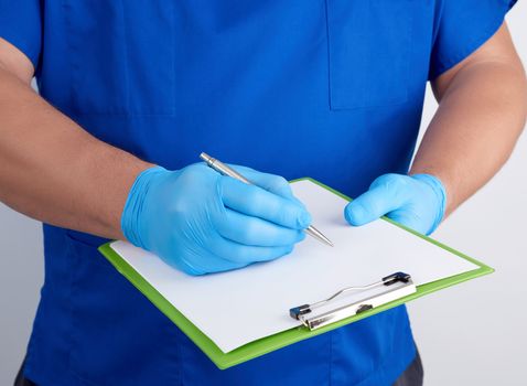 doctor in blue uniform and latex gloves holds a green holder for sheets of paper and writes with his right hand, close up