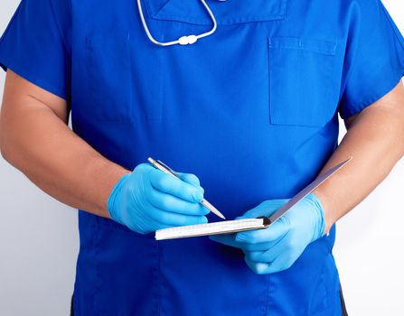 doctor in blue uniform and sterile latex gloves holding open blank notebook, white background, writes with his right hand