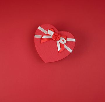closed red gift box in the form of a heart with a bow on a red background, copy space, flat lay