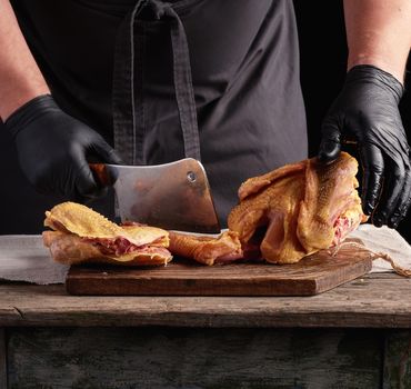 chef in black uniform and latex gloves chopping raw chicken into pieces on a brown wooden board, dark background, cooking process