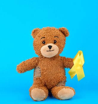 brown teddy bear sits and holds in his paw a yellow silk ribbon on a blue background, concept of the fight against childhood cancer. problem of suicides and their prevention