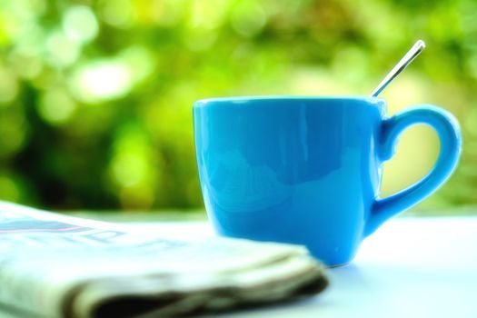 Selective and soft focus of blue coffee cup with blurred newspaper and natural green background