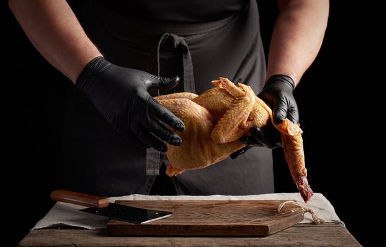 chef in black latex gloves holds a whole chicken carcass over a brown cutting board, meat cooking process, dark background