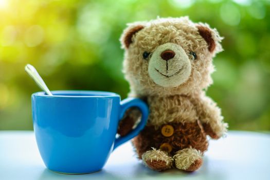 Brown bear doll sitting with a cup of coffee on blurred green natural background