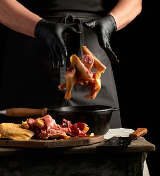 chef in black uniform and latex gloves chopping throws sliced chicken into a black cast-iron frying pan with a wooden handle, meat flies into a container, dark background