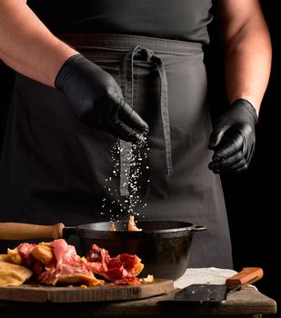 chef in black uniform and latex gloves sprinkles with white salt raw chicken meat in a black cast-iron frying pan, cooking