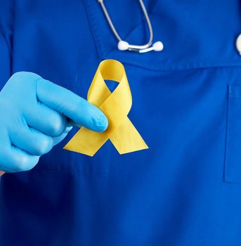 doctor in a blue uniform and sterile latex gloves holds a yellow ribbon in his hand, symbol of the fight against the cat's tumor, problem of suicides and their prevention, close up