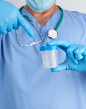 doctor in blue uniform and latex gloves is holding an empty plastic container for taking urine samples, close up