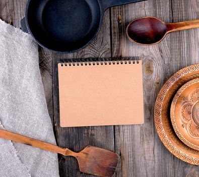 empty black round frying pan with handle, spoon and paper notebook on a wooden table, top view