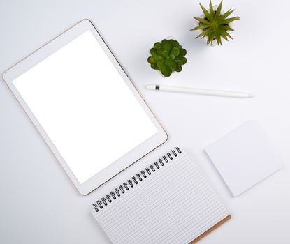 white electronic tablet with a blank screen and a pencil, near pots with green plants on a white table, top view, designer workplace