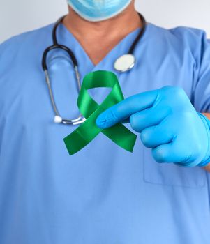 doctor in blue uniform and latex gloves holds a green ribbon as a symbol of early research and disease control,  symbol of Lyme disease, kidney transplantation and organ donation