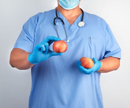 doctor in blue uniform and sterile latex gloves holds ripe red apples, healthy eating concept, white background