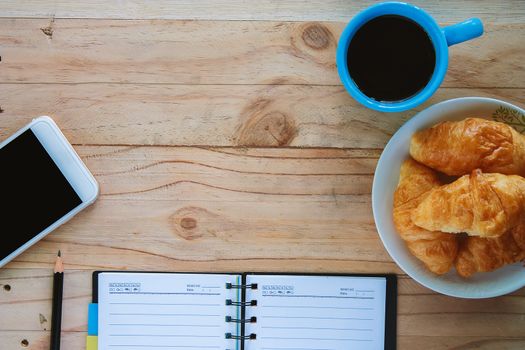 Top view of notebook, a cup of coffee, mini croissant bread, mobile smartphone,and pencil on the wooden table, space for thinking and writing with the snack time