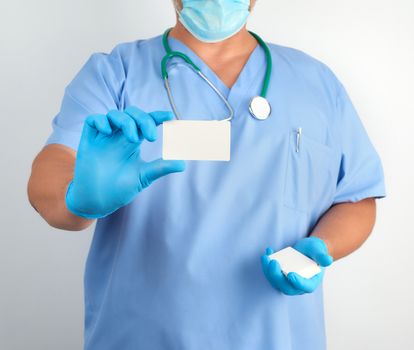 doctor in sterile latex gloves and blue uniform holds a blank white business card, template for the inscription of contacts