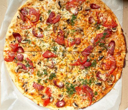 baked round pizza with smoked sausages, mushrooms, tomatoes, cheese and dill in an open cardboard box on a wooden table, food is sliced in portions, top view