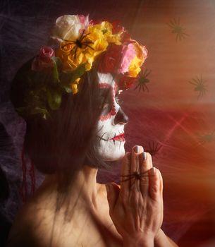 girl with black hair is dressed in a wreath of multi-colored roses and makeup is made on her face Sugar skull to the day of the Dead, her hands are folded in a prayer gesture