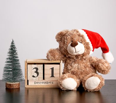 brown teddy bear in red hat, desk wooden calendar with the date december 31 and green decorative tree, christmas background