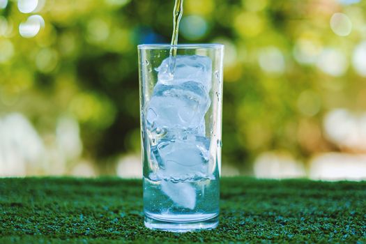 Pouring water into a glass of ice with blurred natural green background and added color filter