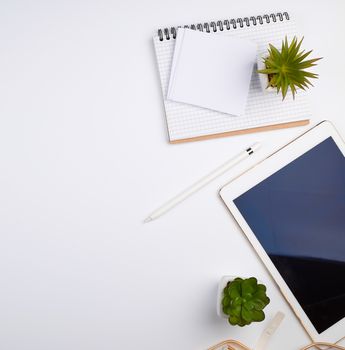 white electronic tablet with a blank screen and a pencil, near pots with green plants on a white table, top view, designer workplace