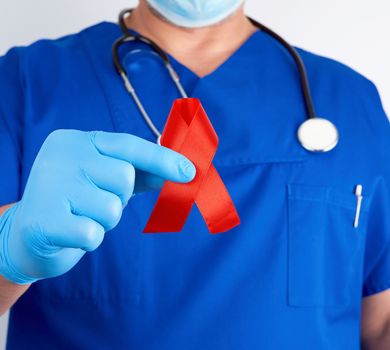 doctor in blue uniform and sterile latex gloves holds a red ribbon-symbol of the fight against disease AIDS and vasculitis, close up