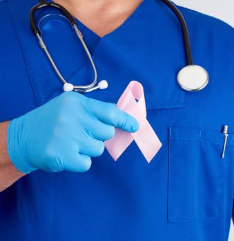 doctor in blue uniform and sterile latex gloves holds a pink ribbon a symbol of the fight against breast cancer in October every 20, close up