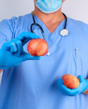 doctor in blue uniform and sterile latex gloves holds ripe red apples, healthy eating concept, white background, close up