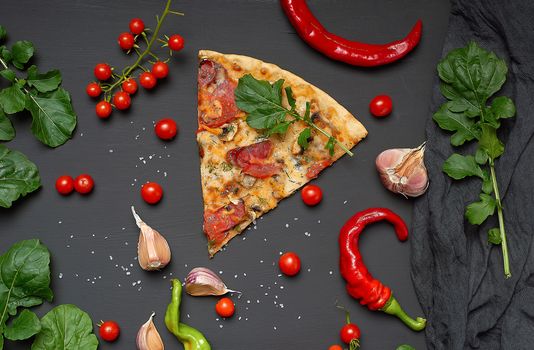 triangular slice of baked pizza with mushrooms, smoked sausages, tomatoes and cheese, next to fresh green leaves of arugula, black background, flat lay