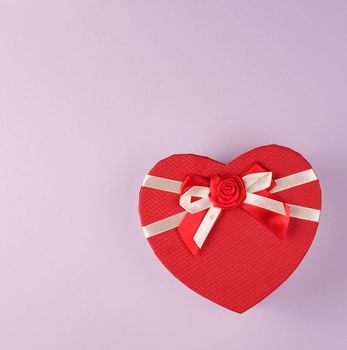 closed red gift box in the form of a heart with a bow on a purple background, copy space