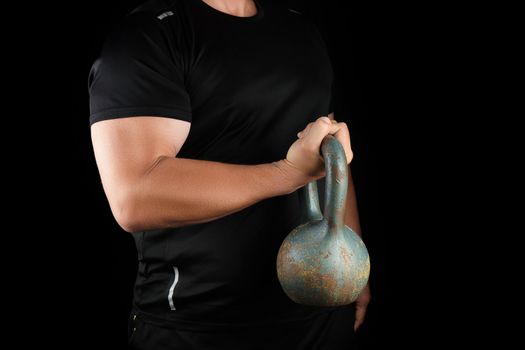 adult strong athlete in black clothes holding an iron kettlebell on his outstretched arms, black background