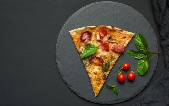 delicious triangular slice of pizza with smoked sausages, mushrooms, tomatoes, cheese and basil leaves on a black graphite board, top view, copy space