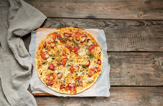 baked round pizza with smoked sausages, mushrooms, tomatoes, cheese and arugula leaves, food is cut in portions, wooden table, copy space