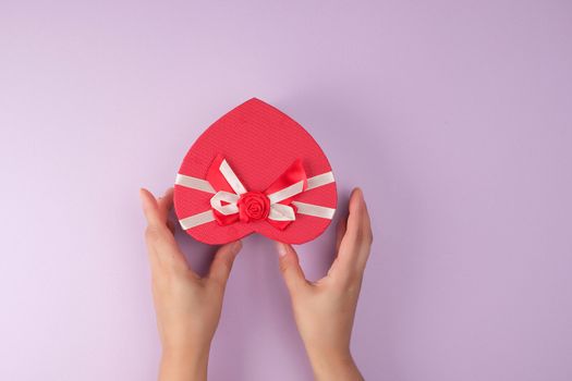 two hands hold a paper closed red box in the shape of a heart, festive backdrop