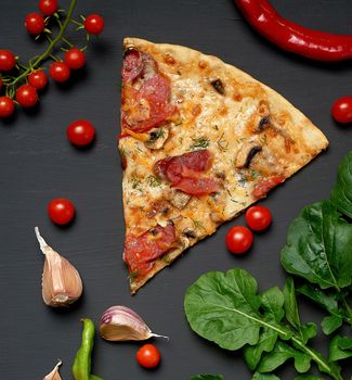 triangular slice of baked pizza with mushrooms, smoked sausages, tomatoes and cheese, next to fresh green leaves of arugula, black background, flat lay