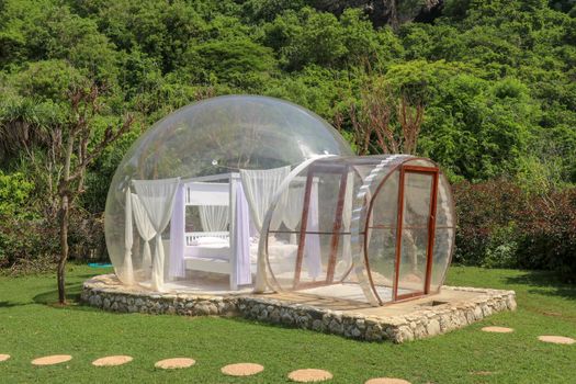 Glamping on Bali beach, in bubble house with transparent walls. White wooden double bed with mosquito net inside bubble. Honeymoon in an inflatable tent. Tourist attraction in tropical paradise.