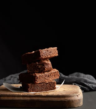 stack of baked square pieces of chocolate brownie cake on brown wooden cutting board, black background