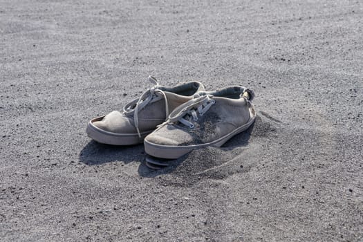 Old torn white sneakers on black volcanic sand. A pair of tie shoes with laces in volcanic ash. Discarded destroyed shoes.