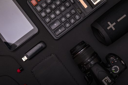 Top view of pitch black office desk with calculator, usb drive, earphones, dslr camera and wireless speaker. Minimal black design concept.