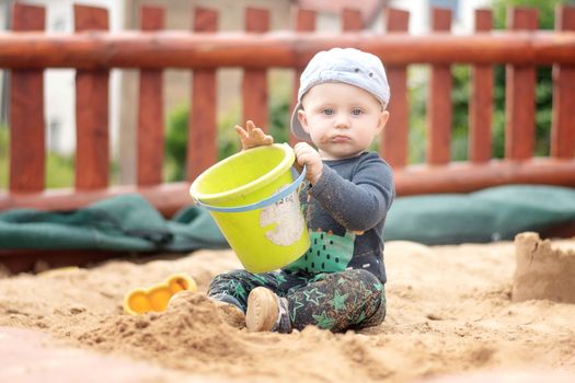 A Caucasian boy in a baseball cap sits in a sandbox with a green bucket in his hand and his mouth dirty from the sand. Looking into the camera. Fun on the kindergarden.