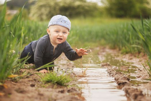 A Caucasian infant in a cap is playing in a muddy pool. Children's joy of freedom. Looking into the camera.
