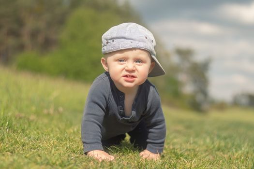 92/5000
An 11-month-old infant in a peaked cap climbs the lawn. Looking into the camera. Blurred background.