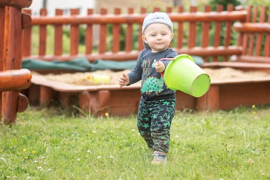 A Caucasian boy in a baseball cap walks across the lawn at the playground, holding a green bucket of sand in his hand. Looking into the camera. An 11 month old boy enjoys a day playing outside.