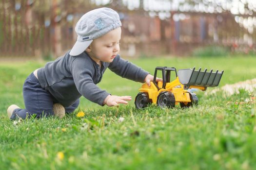 A Caucasian infant in a peaked cap plays on the lawn with an excavator model. A small child with a toy in the garden.