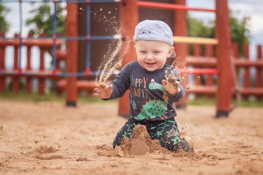 Caucasian boy in baseball cap playing on sand at playground. An 11 month old boy throws sand in the air.