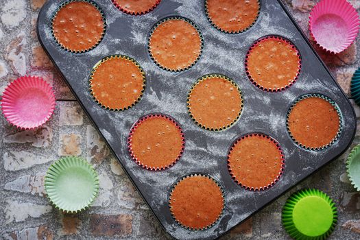 Baking tray for muffins with raw dough on earthenware background surrounded by paper colored baskets. View from above.