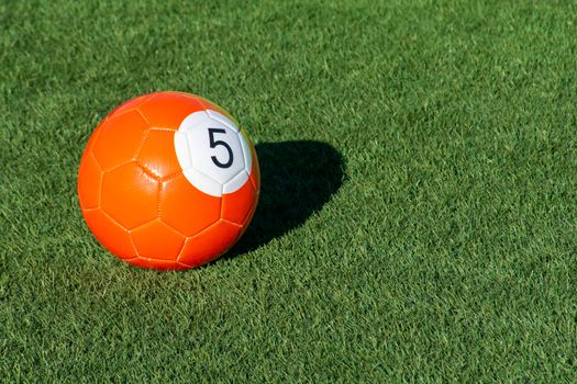 Bright orange number 5 soccer billiards or pool ball on green grass with a shadow and copy space. Concept of sports, recreation and childhood fun.