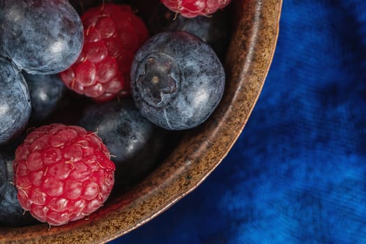 Clay bowl with raspberries and blueberries on a blue scarf. Close up. Top view.