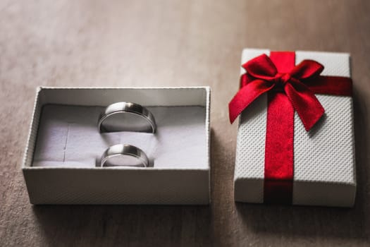 Marry me concept. White box with two engagement rings and red ribbon on a wooden table.