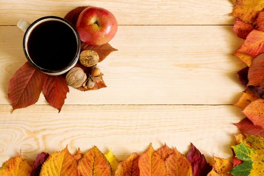 A tin cup with coffee on a wooden table with a frame of fallen colorful leaves, with apple and nuts.