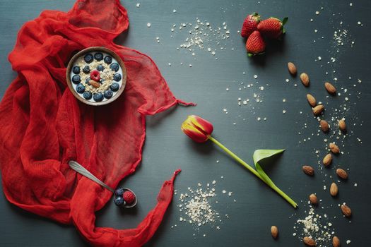 Breakfast with love. Bowl of oatmeal with fruit and almonds on a gray table with tulip and red scarf.